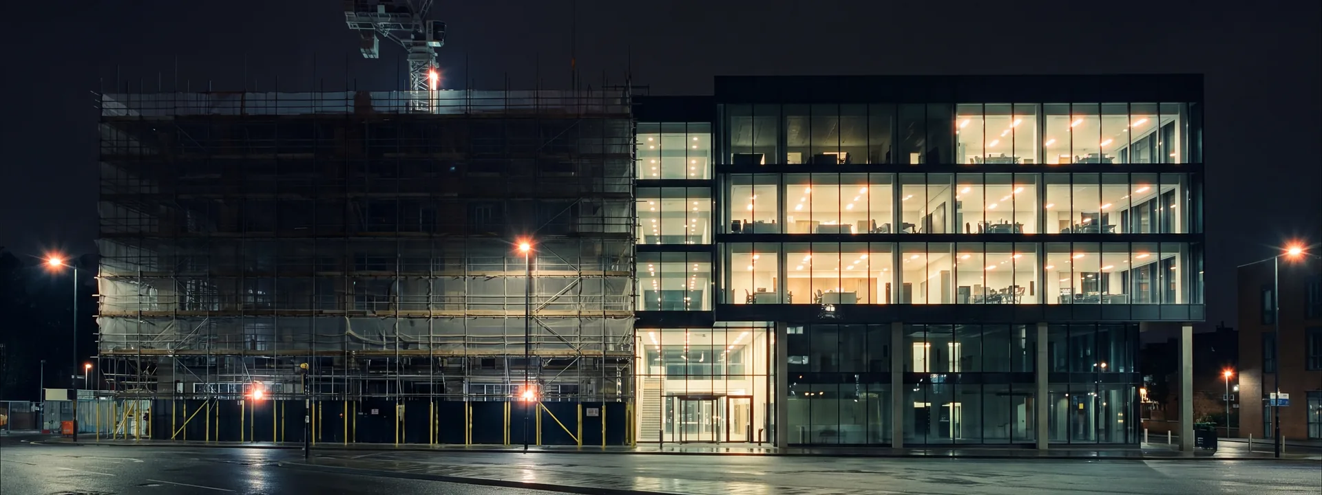 One office building at night: scaffold and a tower crane on the left, the finished glazed floors lit and occupied on the right